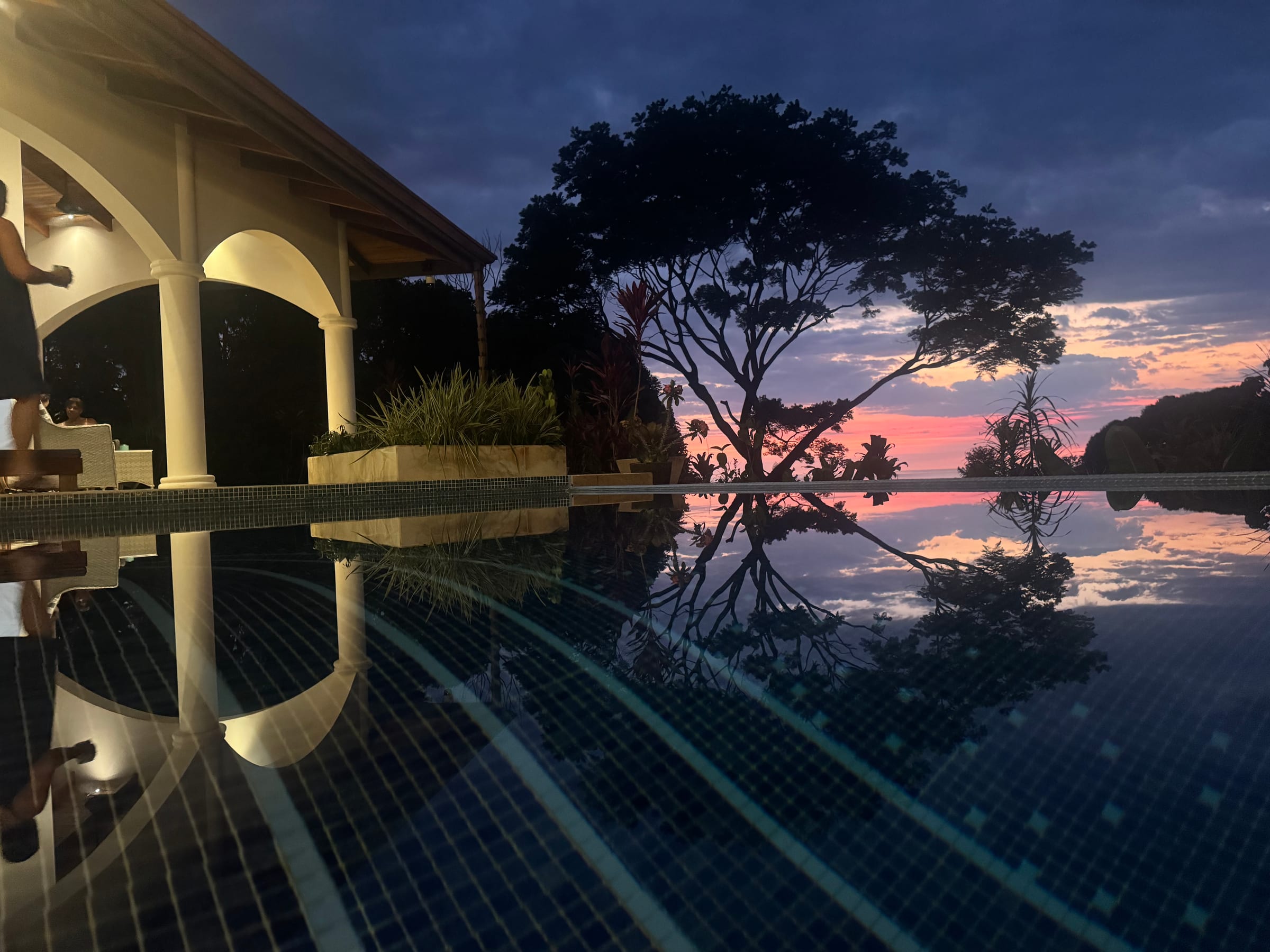 Sunset reflected in the infinity pool at Casa Rosa Blanca, the villa's lit arches on the left and a tree silhouetted against a pink sky above the Pacific, Ojochal, Costa Rica